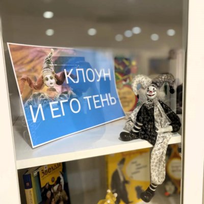 В стеклянной витрине сидит тряпичный клоун и стоит заголовок выставки